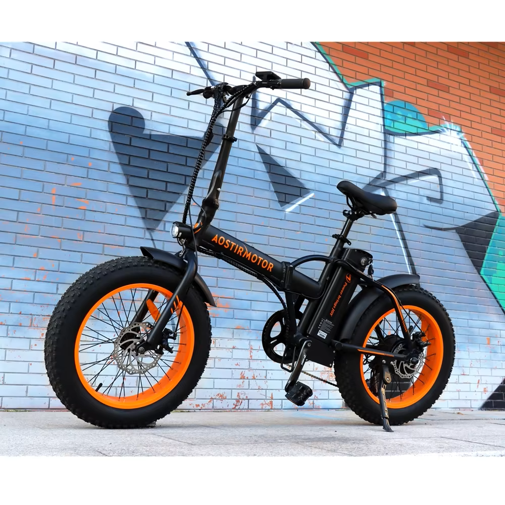 AOSTIRMOTOR Folding Electric Mountain Bike with bold orange fat tires, sturdy black frame, and sleek design, parked against a graffiti-covered urban wall.